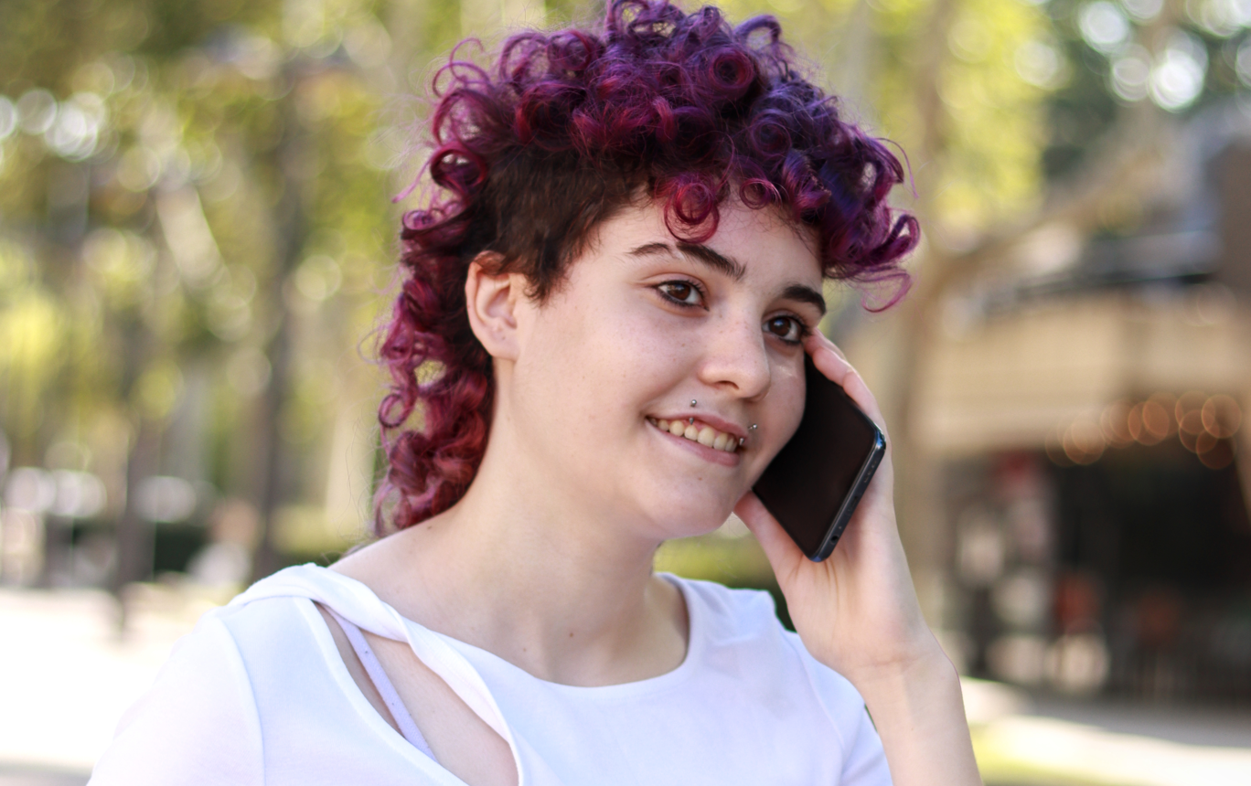 Person with purple-tinged curly hair wearing a white tshirt with a smart phone held to their ear.